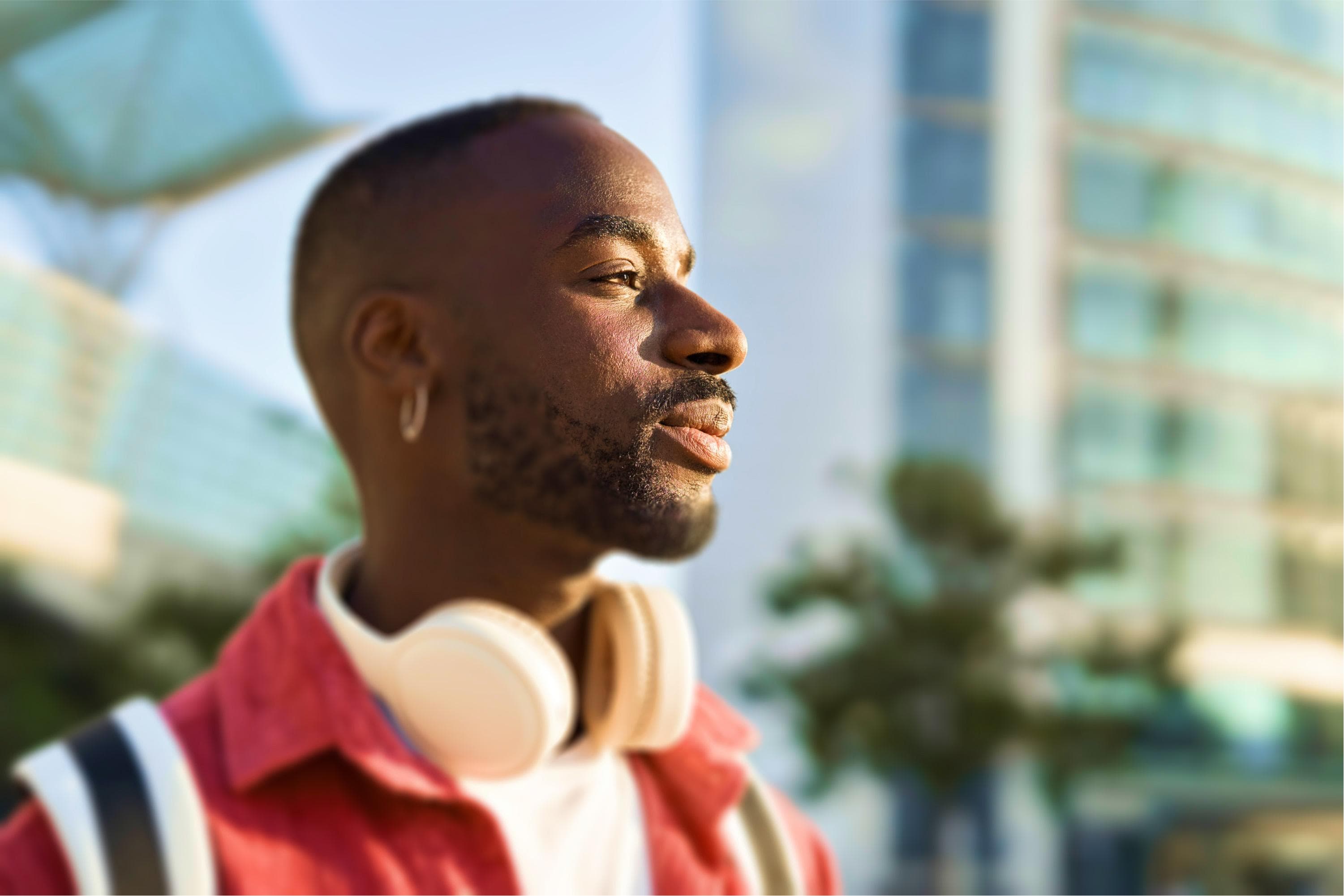 A close-up side view of a person wearing headphones around their neck. They are smiling gently and dressed in a casual red jacket, standing outdoors with skyscrapers and trees in the soft-focus background.