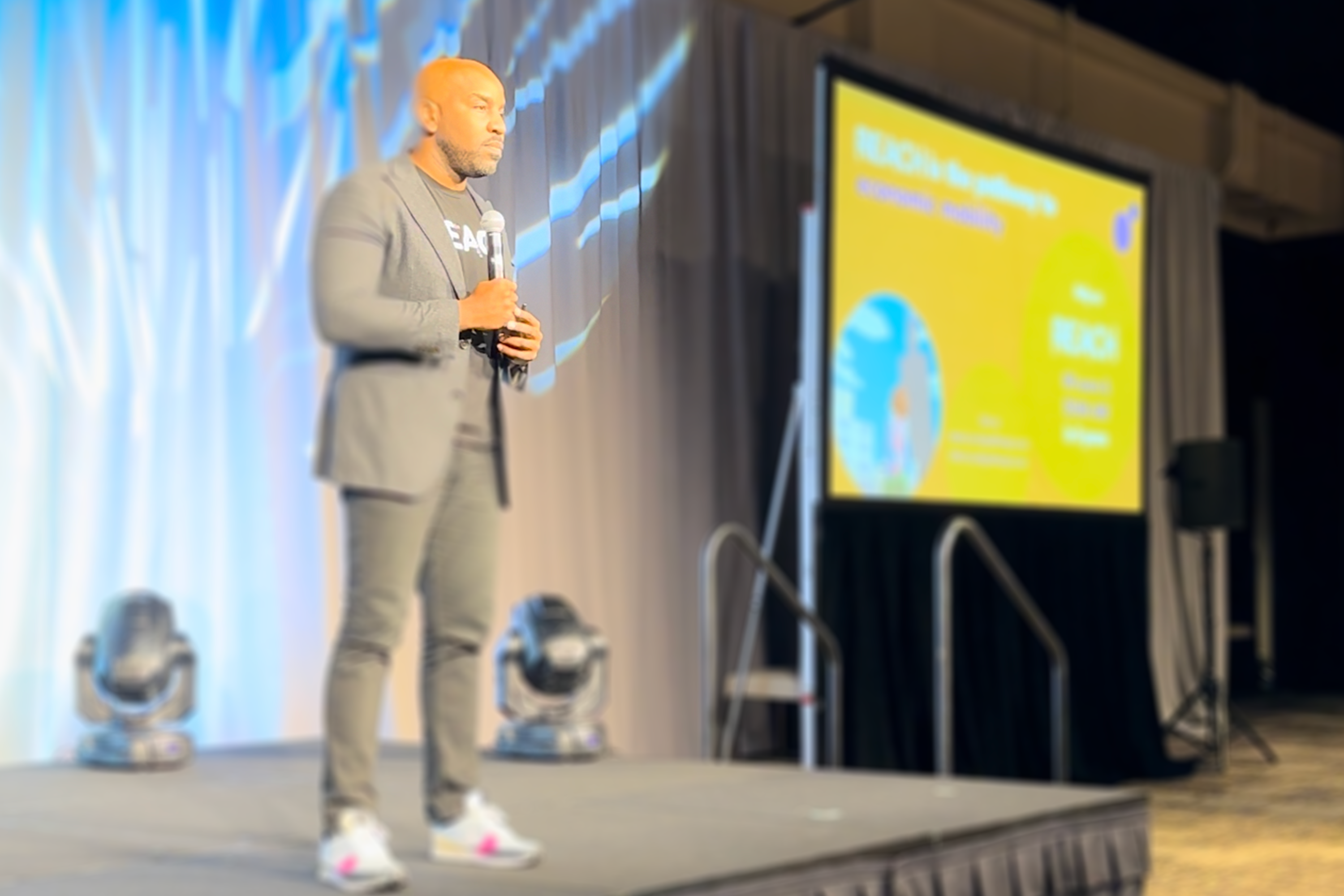 Jeffery Beckham Jr. stands on stage at a conference, dressed in a casual grey blazer and trousers with a grey t-shirt, holding a microphone. In the background, there is a large screen displaying a presentation for Reach Pathways.