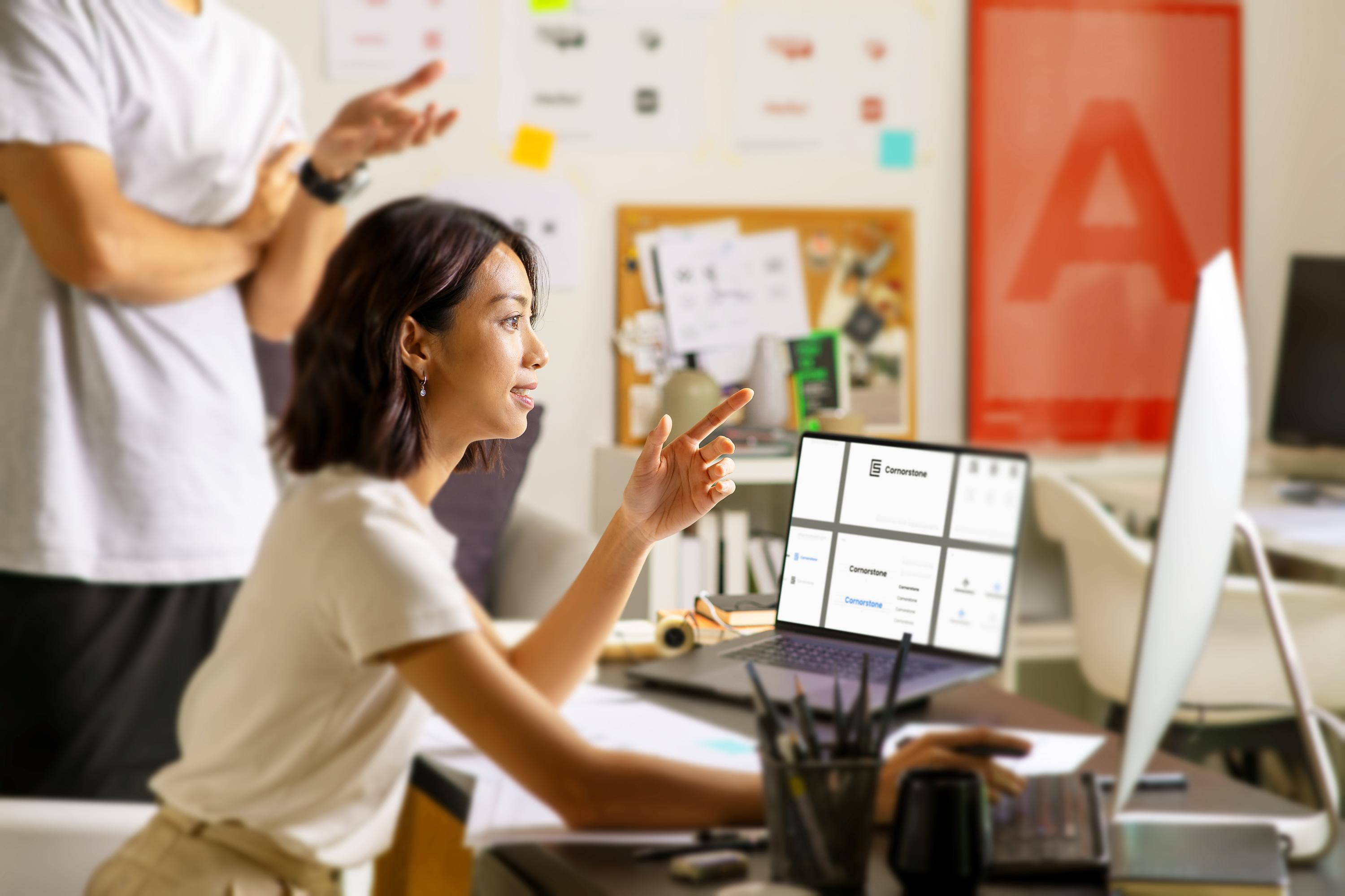 In a creative office setting, a person gestures expressively during a discussion, pointing towards a computer monitor displaying an innovative user interface.