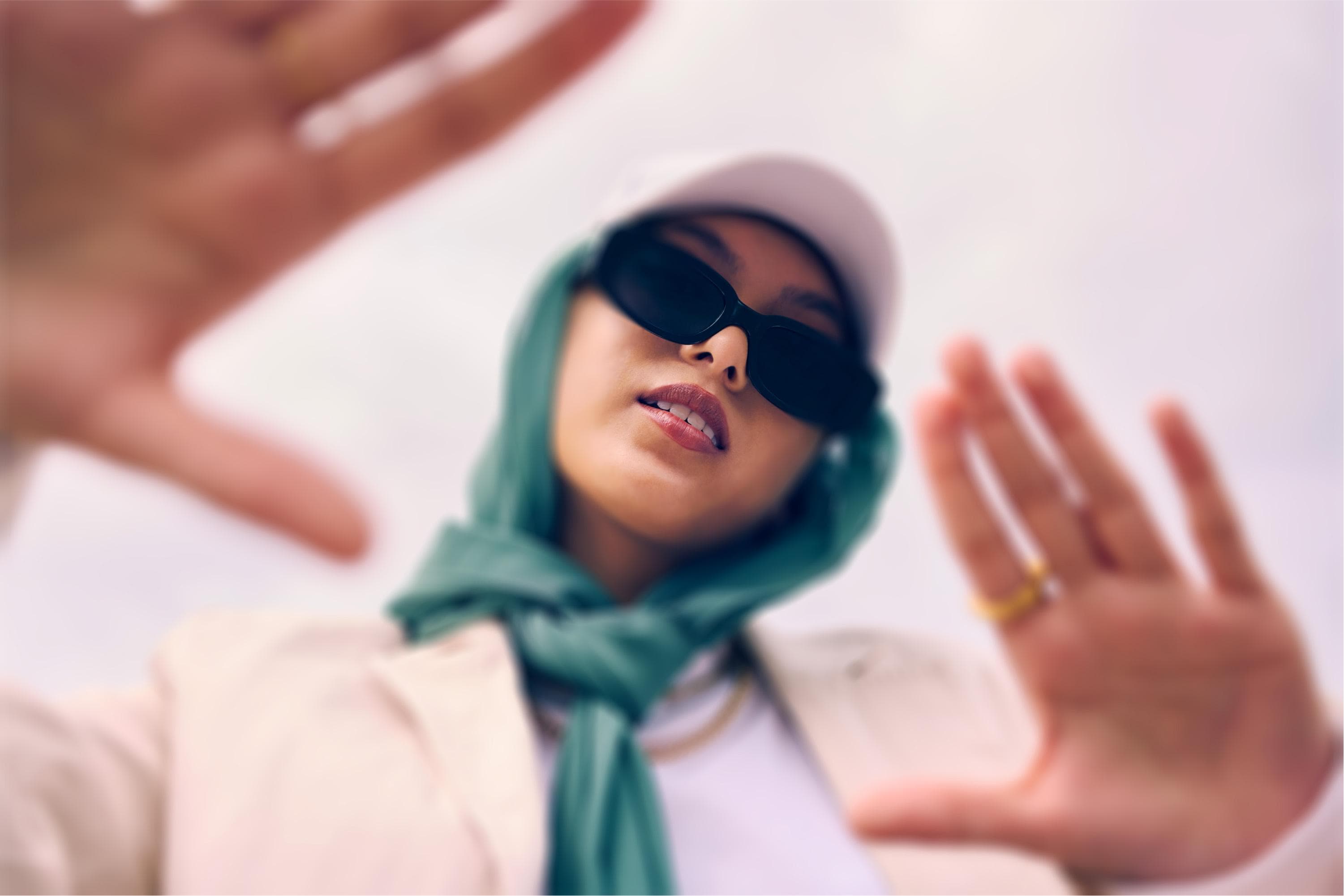 A close-up of a person wearing large dark sunglasses and a light green scarf around their head. They are holding their hands up as if framing a shot, set against a bright sky.