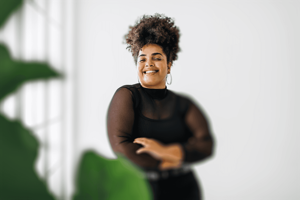 A cheerful individual with curly hair and a black mesh top laughs joyfully in an indoor setting, surrounded by soft natural light.