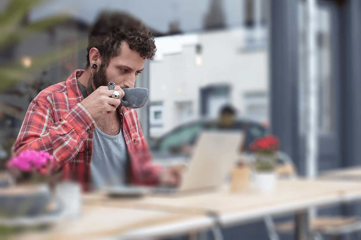 A bearded person sitting at an outdoor café, deeply focused on their laptop screen while sipping coffee from a grey cup.