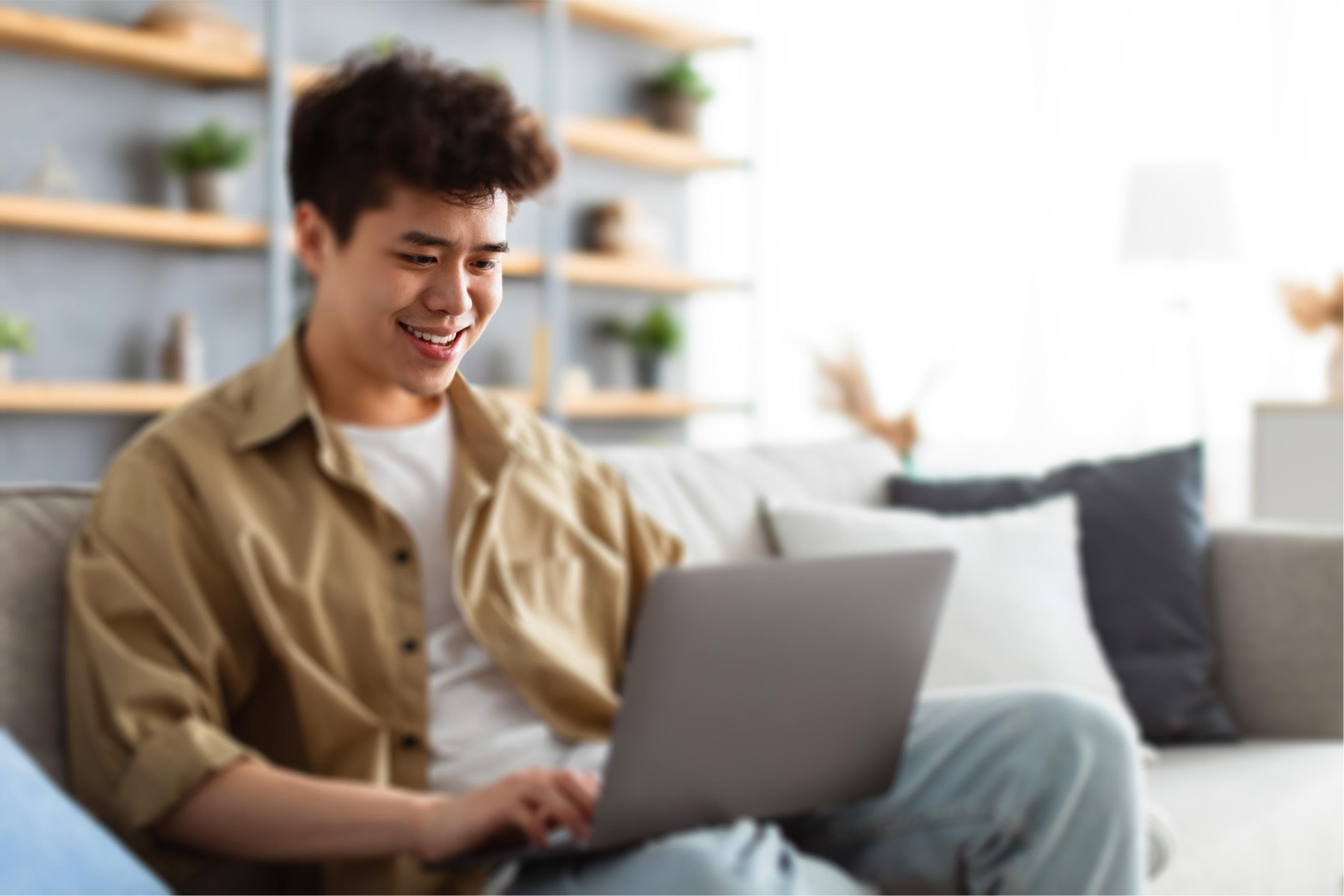 A young person sitting on a sofa, smiling while using a laptop. They are wearing a beige shirt and jeans in a cozy living room setting with shelves filled with plants and decorative items.