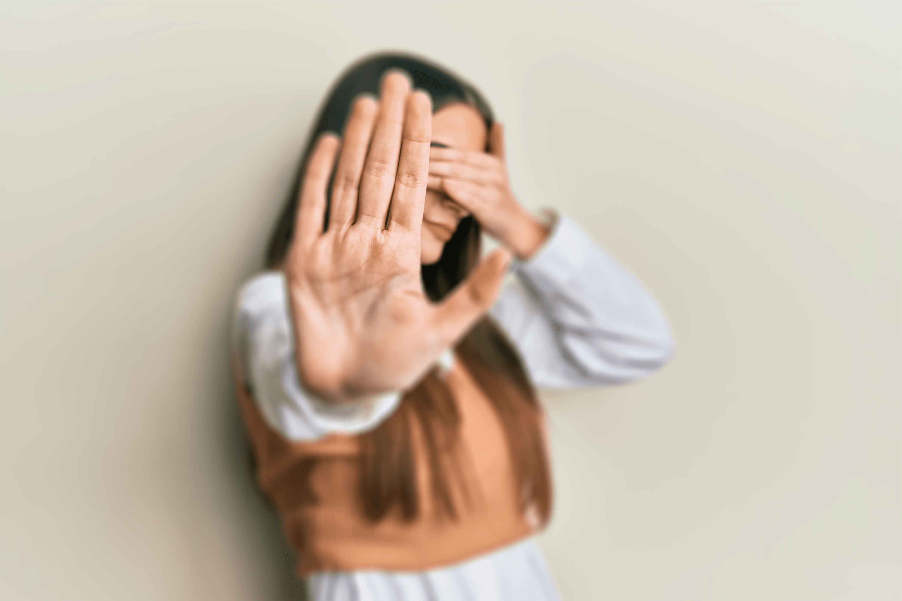 A person with long straight hair, standing against a plain background, covering their face with one hand, while the other hand is placed in front of them in a stop gesture.