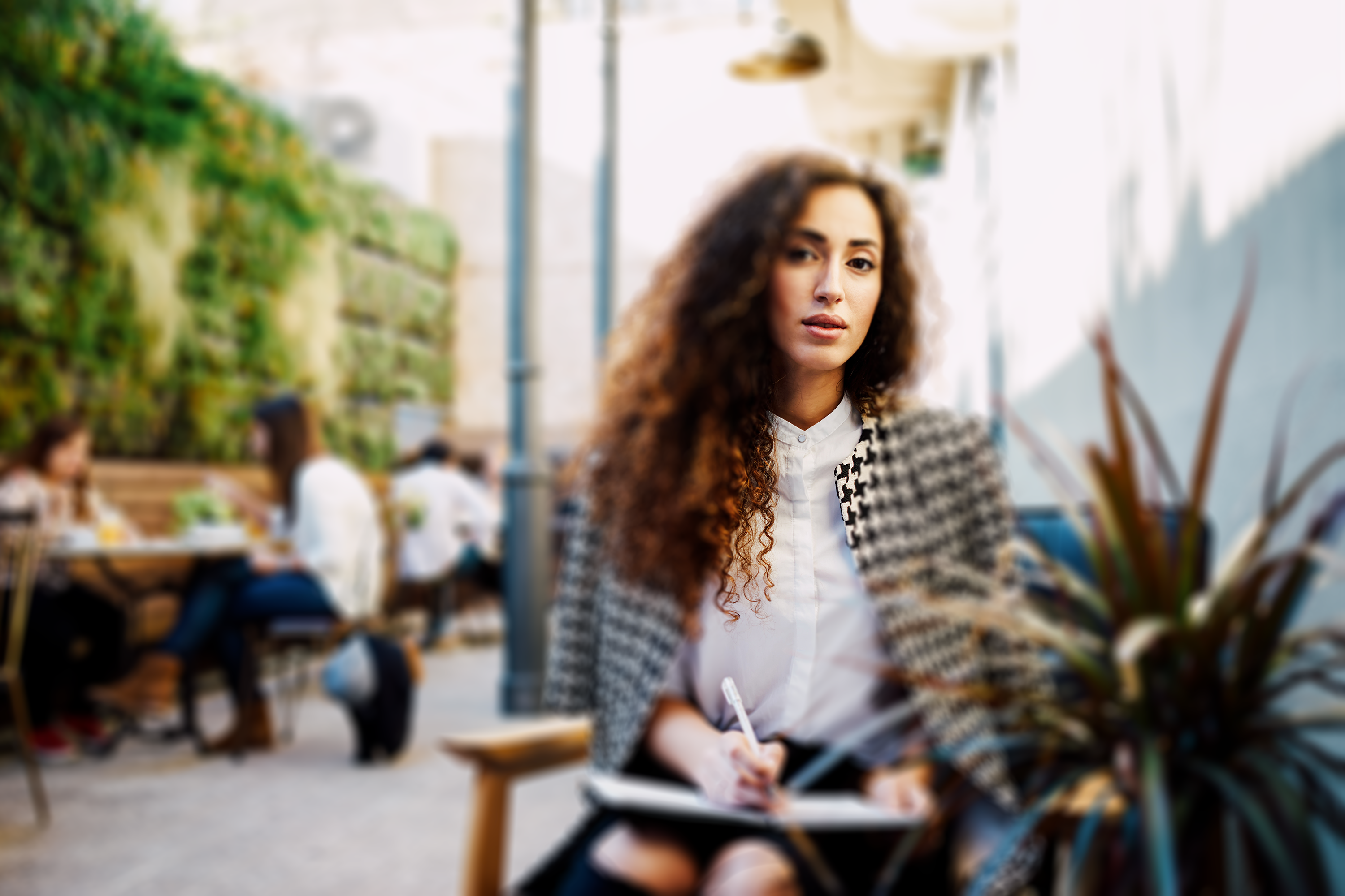 A person with voluminous curly hair, sitting outdoors at a cafe table, looking thoughtful while holding a pen and notebook.