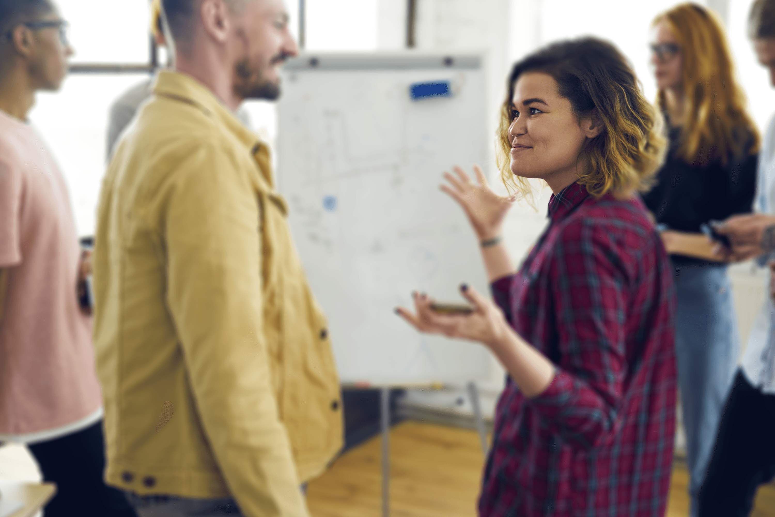 Two people engaging in a lively conversation in a busy office environment. A person in a yellow jacket is smiling at a colleague in a red plaid shirt who is gesturing expressively while holding a smartphone. Behind them, several other colleagues are focused on their devices, standing near a whiteboard filled with business strategies.