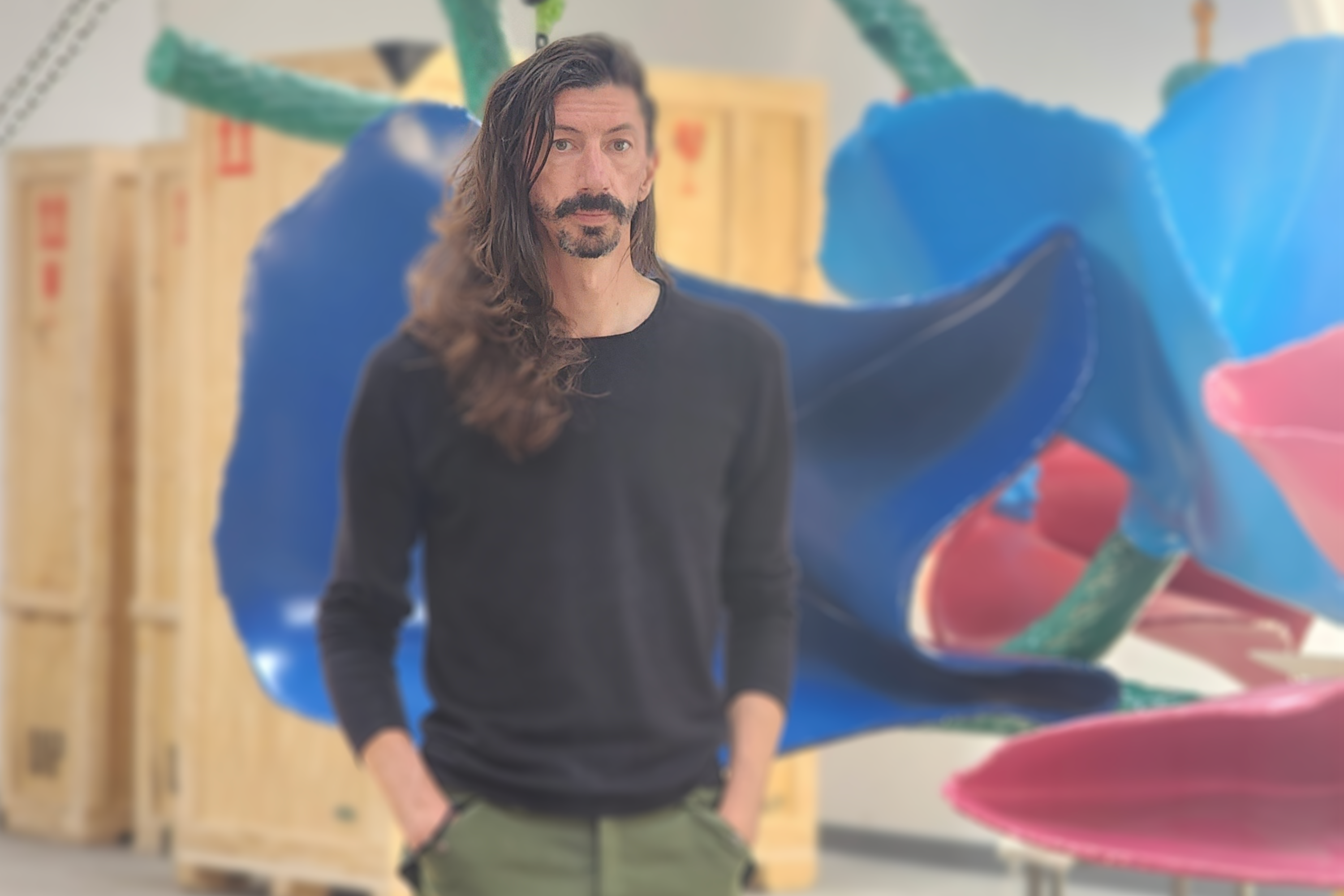 Artist, Sam Stubblefield is featured with long, flowing dark hair and a light beard, dressed in a black long-sleeve shirt and green pants, standing in an industrial-looking space. Behind him is a large, partially visible, colorful abstract sculpture, consisting of vivid blue, red and green elements.