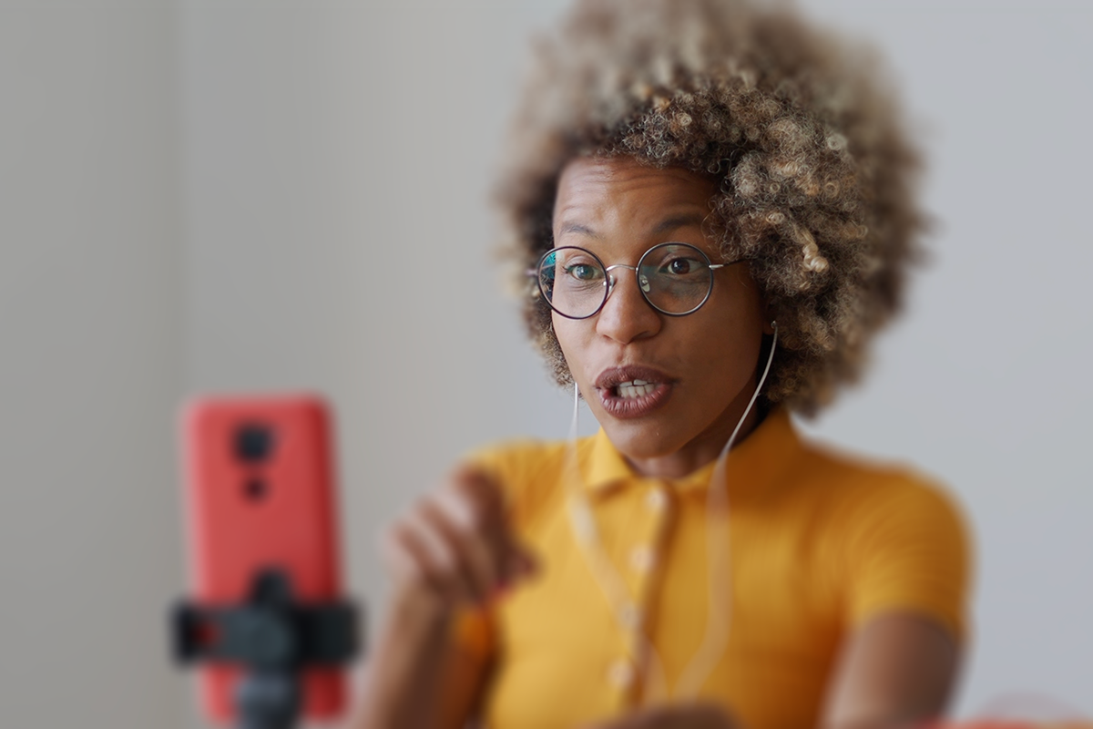 An individual with curly, blonde-highlighted hair and round glasses, wearing a bright yellow shirt. They are recording a video using the red smartphone on a tripod in front of them.