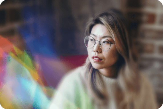 A thoughtful individual with shoulder-length light brown hair and round glasses, looking off to the side with a contemplative expression. They wear a cozy, pastel-colored sweater. The background of the photo is a blurred brick wall