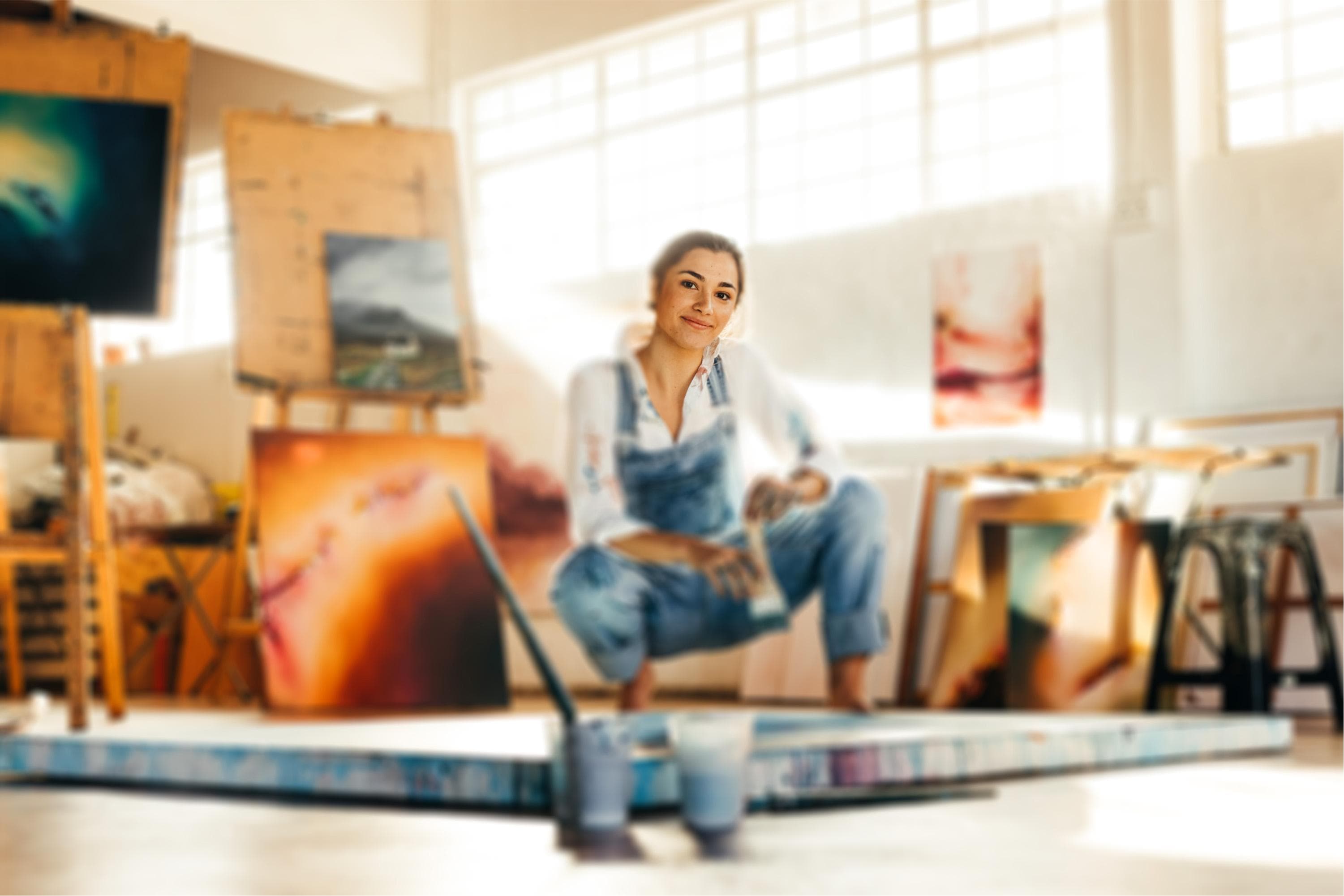 A person in a denim overall sits on a small platform surrounded by vibrant and colorful paintings in a sunlit art studio. The studio is filled with various canvases, some displayed on easels and others propped against the walls. The background shows large windows allowing natural light to enhance the vivid colors of the artwork. 