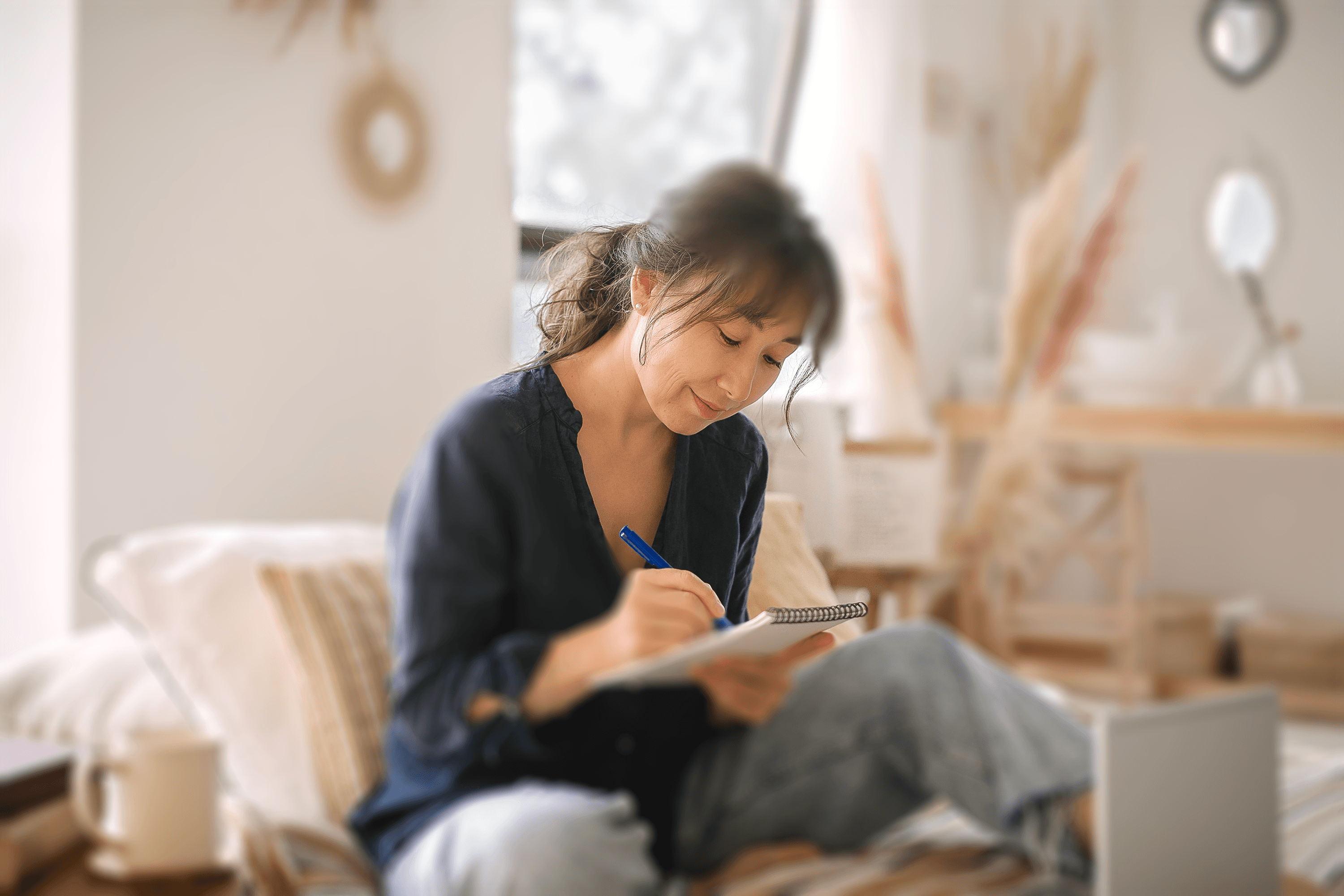 A person with shoulder-length dark hair, seated comfortably on a couch in a cozy, well-lit room, deeply focused on writing in a notebook. They are wearing a casual blue shirt and jeans, and are surrounded by a warm, inviting decor featuring natural tones and soft furnishings. 