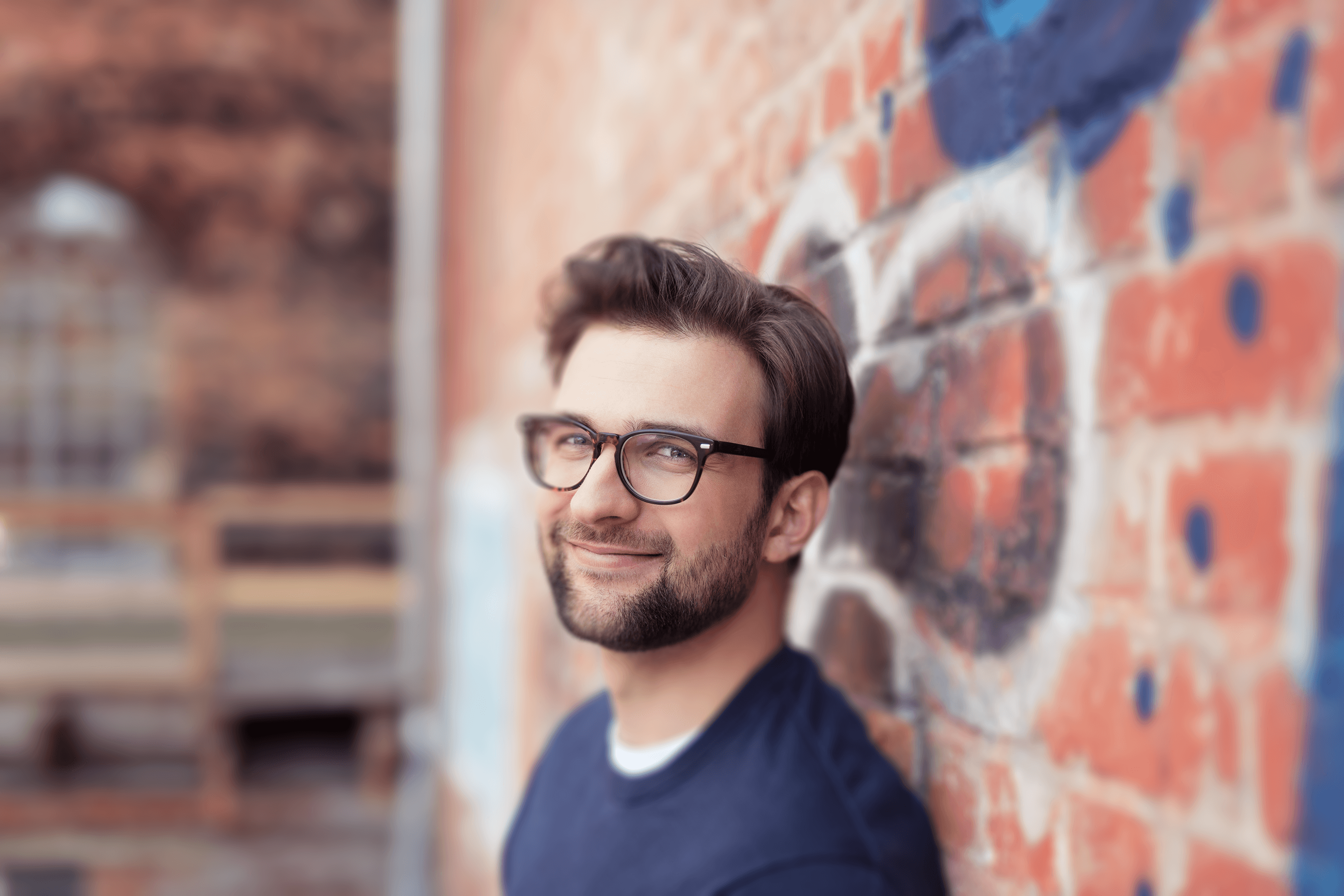 A person wearing glasses and a casual shirt, smiling and looking confident against a brick wall, giving a casual yet professional vibe.