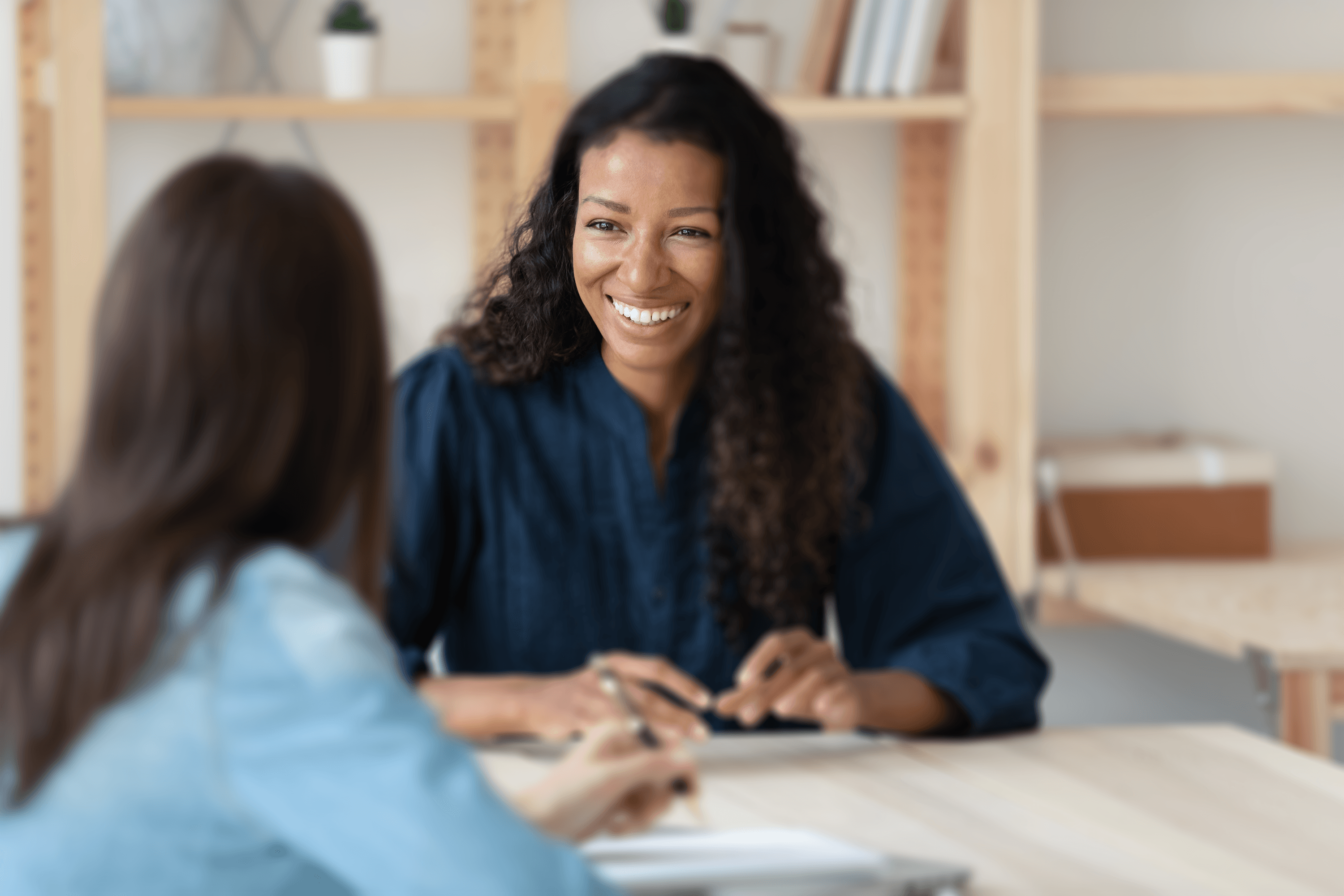 A person with long curly hair, smiling warmly during a meeting at a light wooden table, in a bright office space.