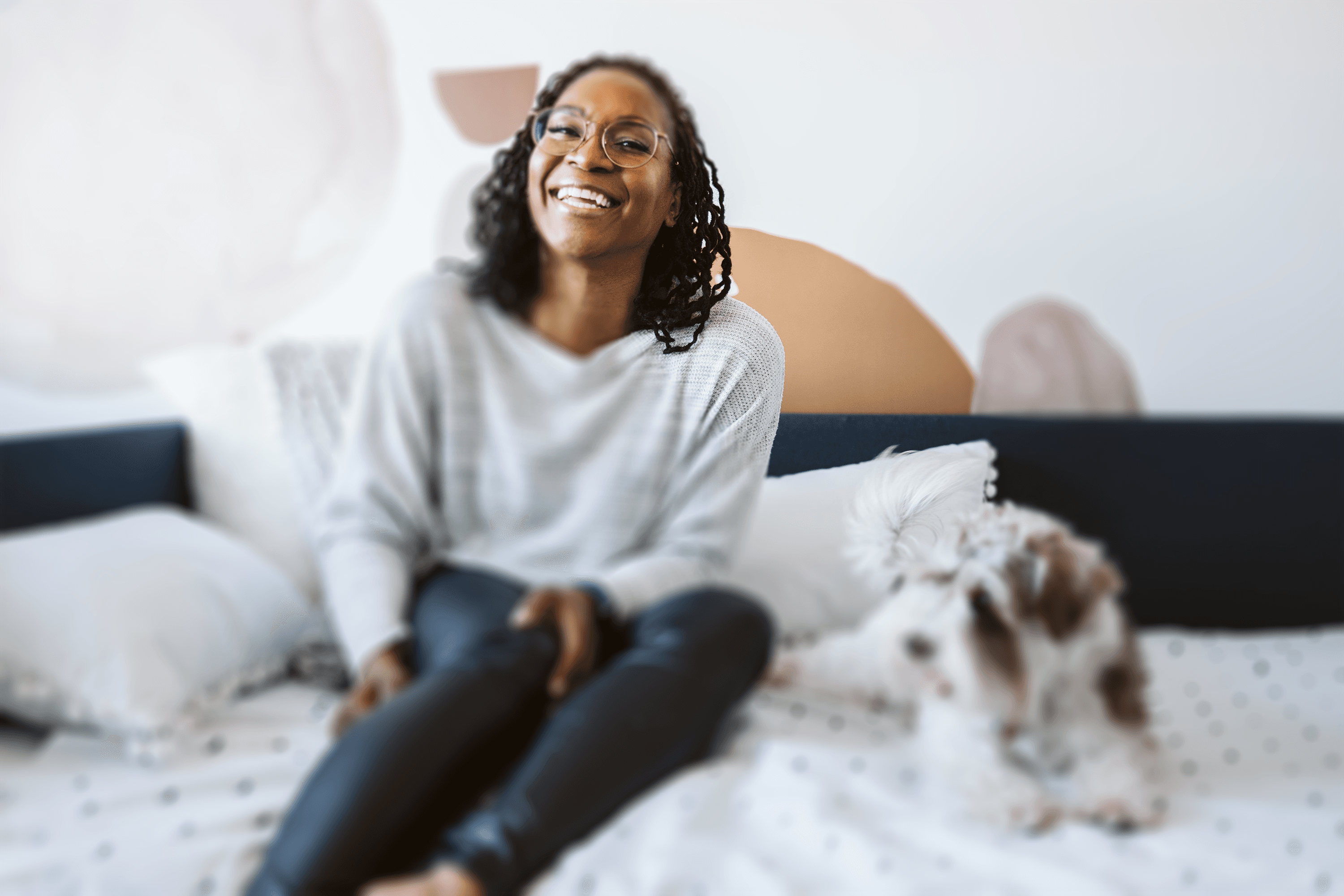 Sharon Milone sits on a bed, smiling at the camera, with her small fluffy dog Ada sitting beside her, in a cozy and stylish bedroom.