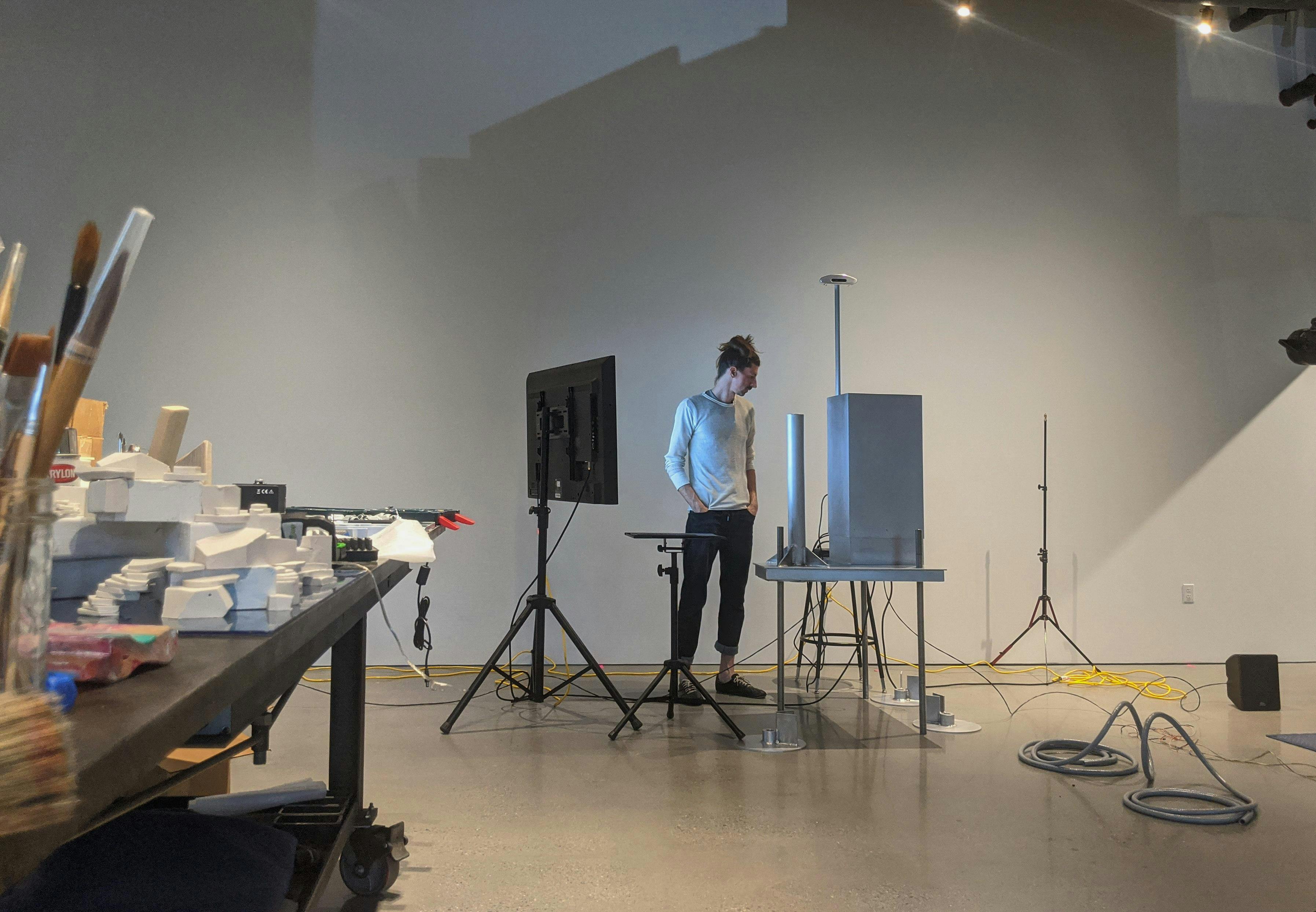 Sam stands focused and engaged in his modern, minimalist art studio. He looks intently at a laptop on a standing desk, surrounded by art materials and technical equipment. The photo embodies the blend of art and technology in his creative process.