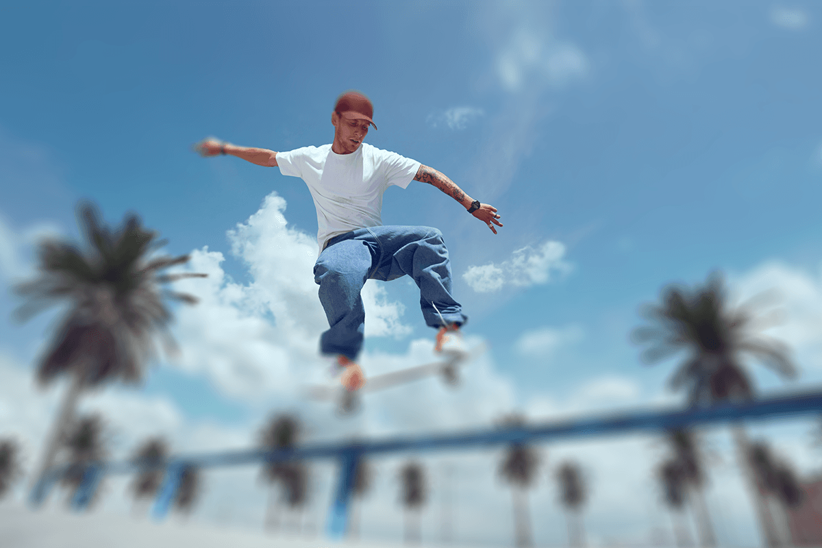 A person in casual attire mid-air while skateboarding, performing a trick against a sunny sky with palm trees in the background.