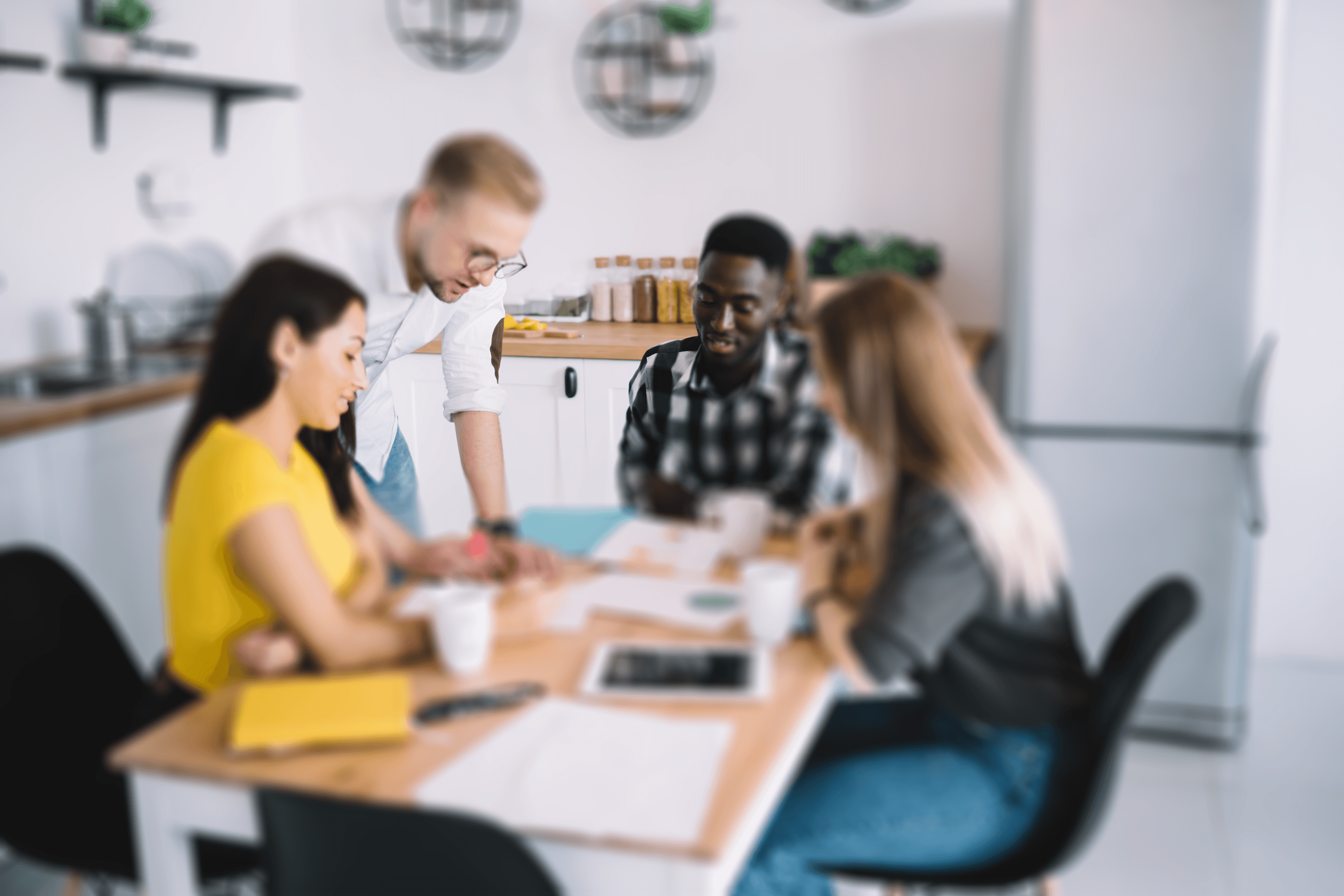 A diverse team of four professionals collaborates around a modern kitchen table in a bright office. They are engaged in an animated discussion, with cups and documents on the table, suggesting a creative brainstorming session.