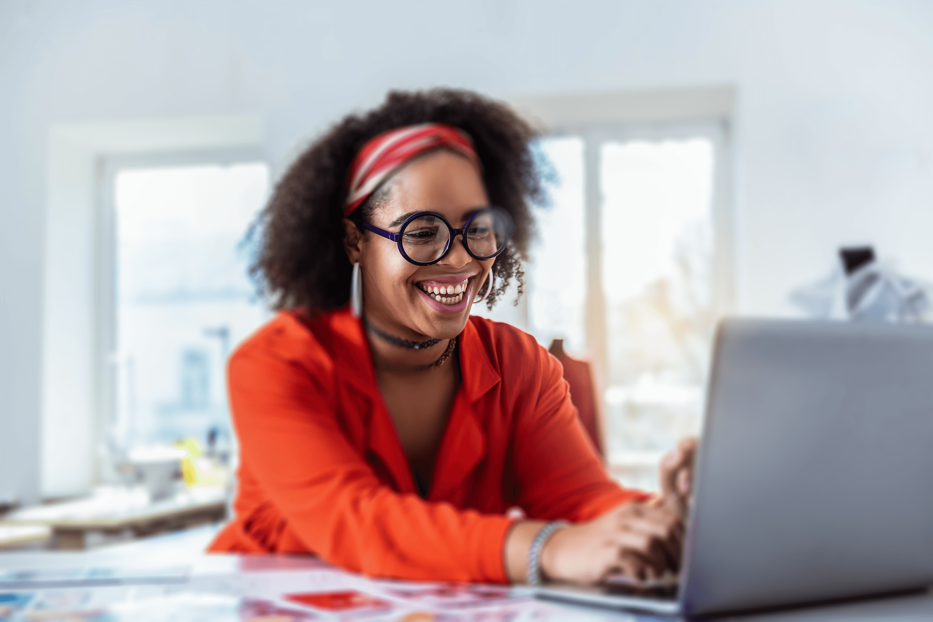 A person with curly hair and glasses, laughing joyfully while looking at their laptop screen in a well-lit room with modern decor.