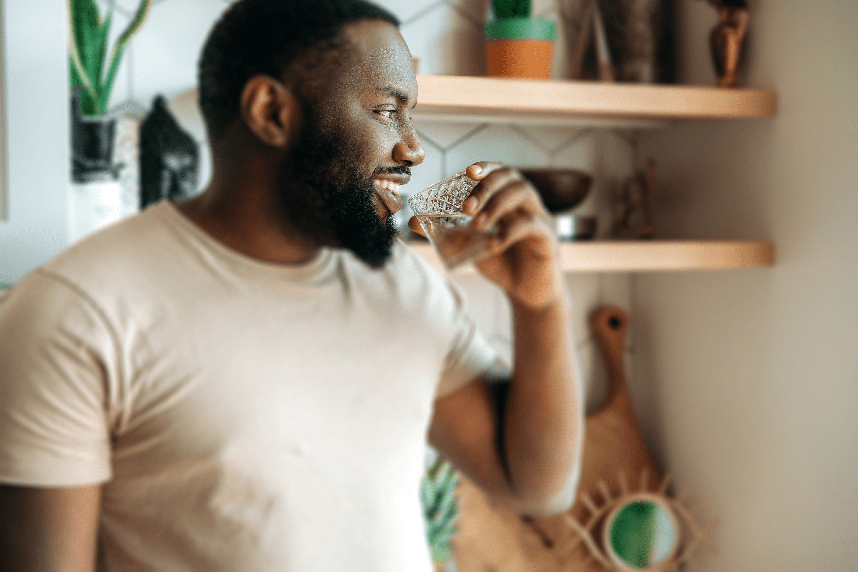 A person in a beige T-shirt, smiling and drinking a small glass of water, stands in a room filled with plants and rustic decor.