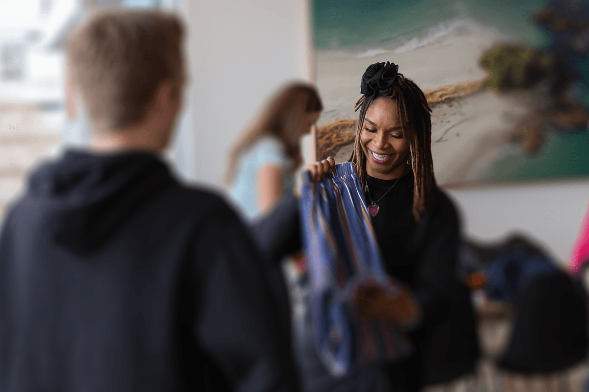 A smiling individual in a black sweater is showing clothing options to another person in a casual office environment, with beachy artwork in the background.