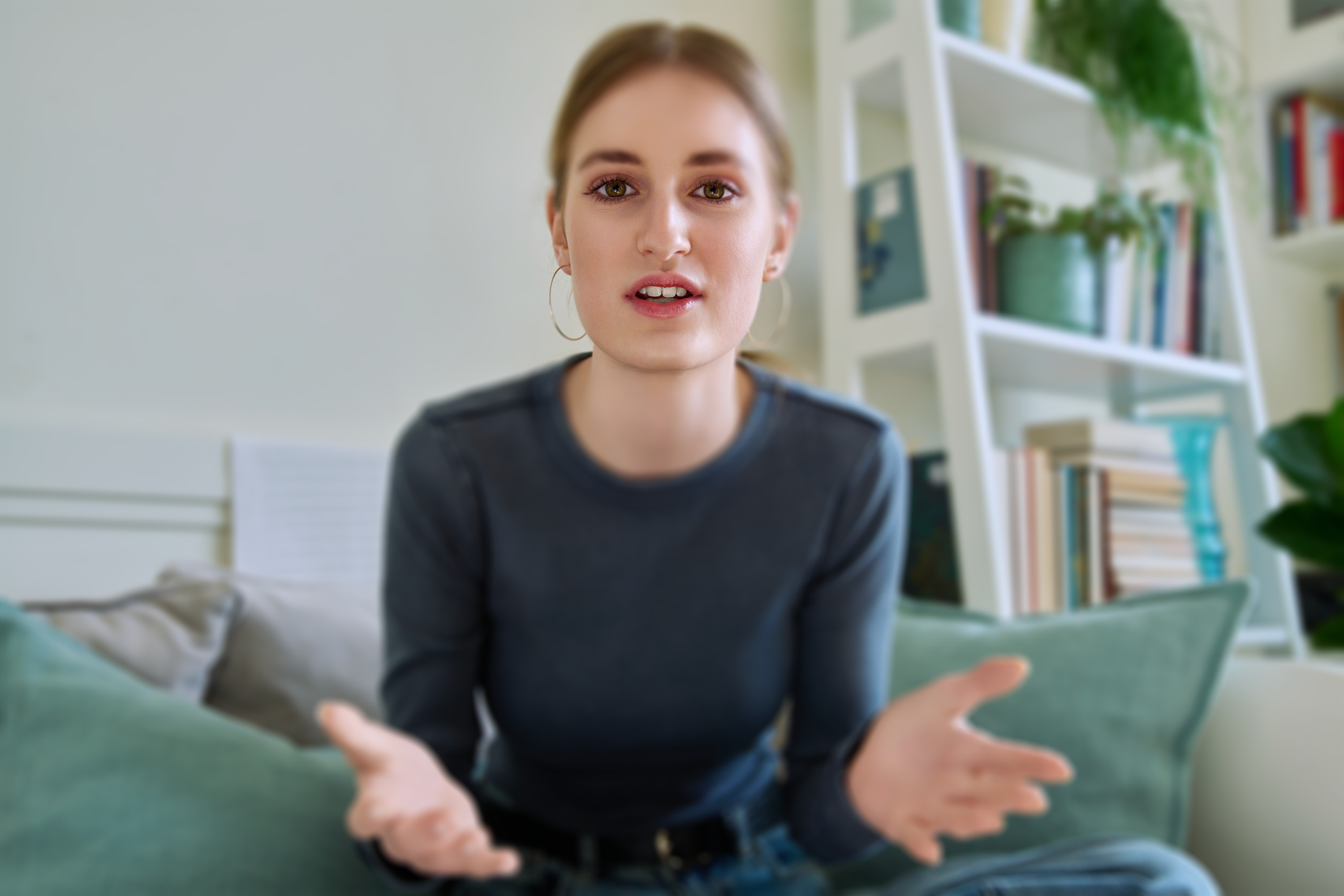 A person with shoulder-length blonde hair and large hoop earrings, wearing a dark blue top. They are seated, gesturing with their hands, and speaking directly to the camera from a cozy room with a modern, well-lit interior featuring a bookshelf filled with books and plants.