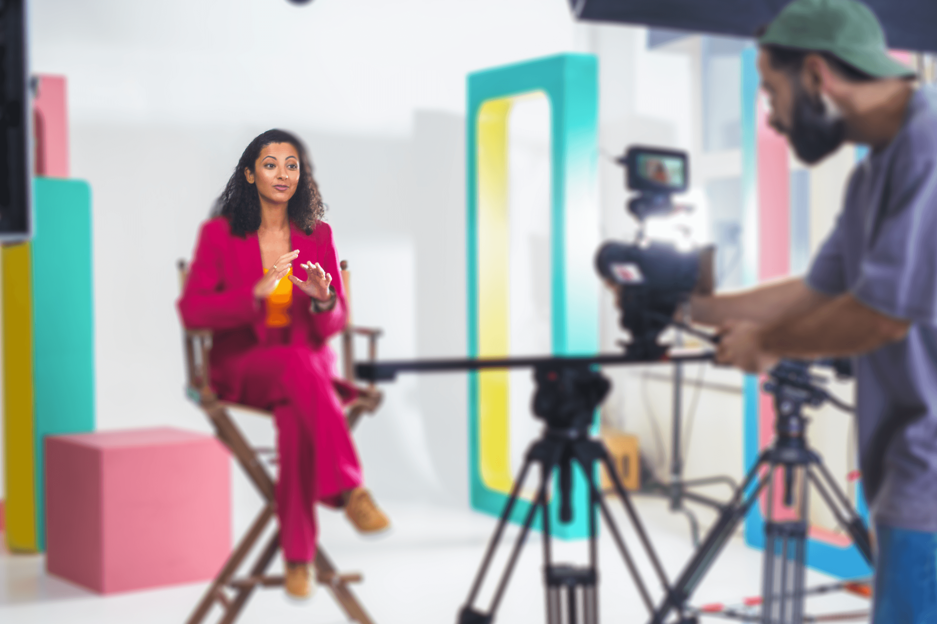 A person in a vibrant magenta suit is seated and speaking animatedly in a colorful video studio set. The scene includes a camera operator filming them, and another camera focused on their face displayed on a monitor. The set features geometric blocks in pastel hues of pink, teal, and yellow, creating a playful and professional backdrop.