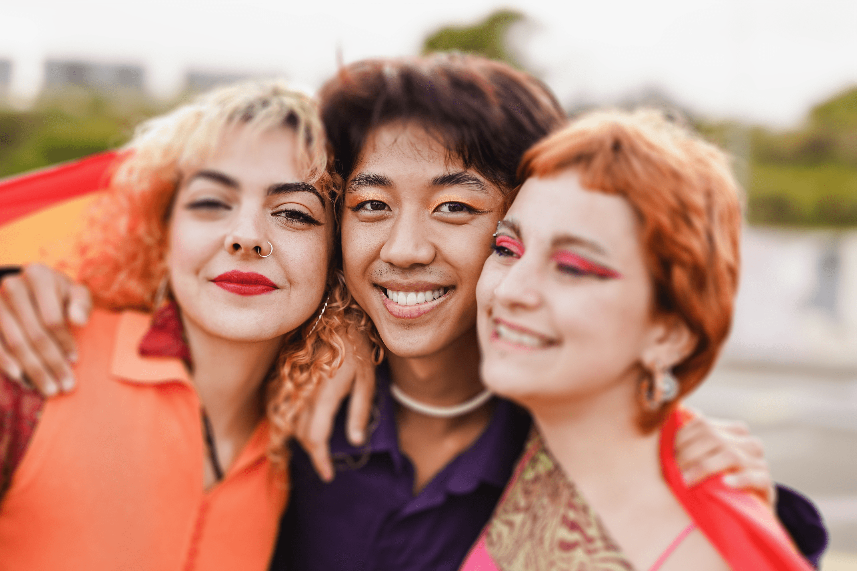 Three joyful friends embrace for a group photo outdoors. On the left, a person with curly blonde hair and a nose ring wears a vibrant orange jacket. In the middle, a person with brown hair and a purple shirt smiles broadly. On the right, a person with curly orange hair and dramatic pink eye makeup grins