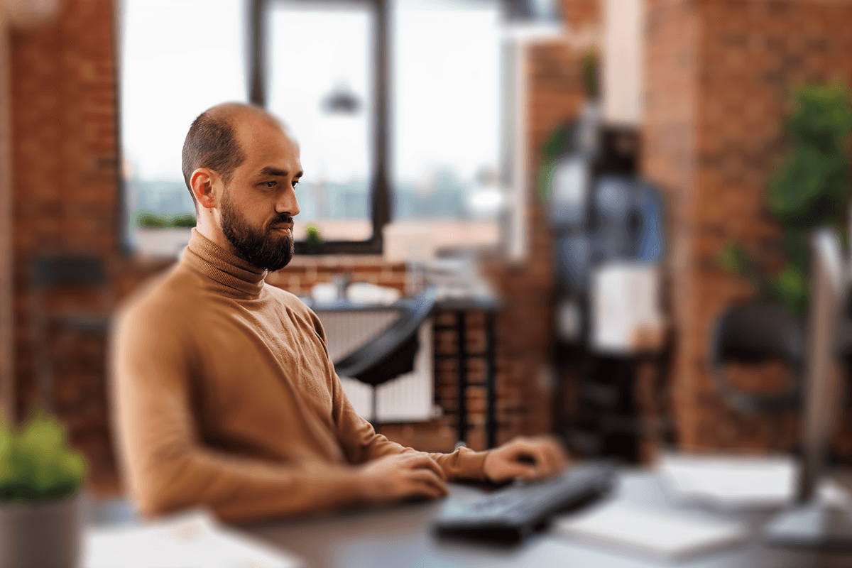 A focused individual with a short beard and shaved head, wearing a turtleneck sweater, seated at a desk and typing. They are in a stylish office with exposed brick walls and large windows.