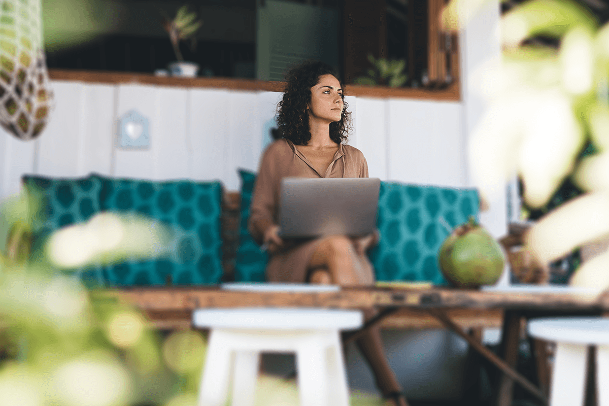 A person sits thoughtfully at an outdoor cafe table, using a laptop in a relaxed tropical setting with vibrant greenery.