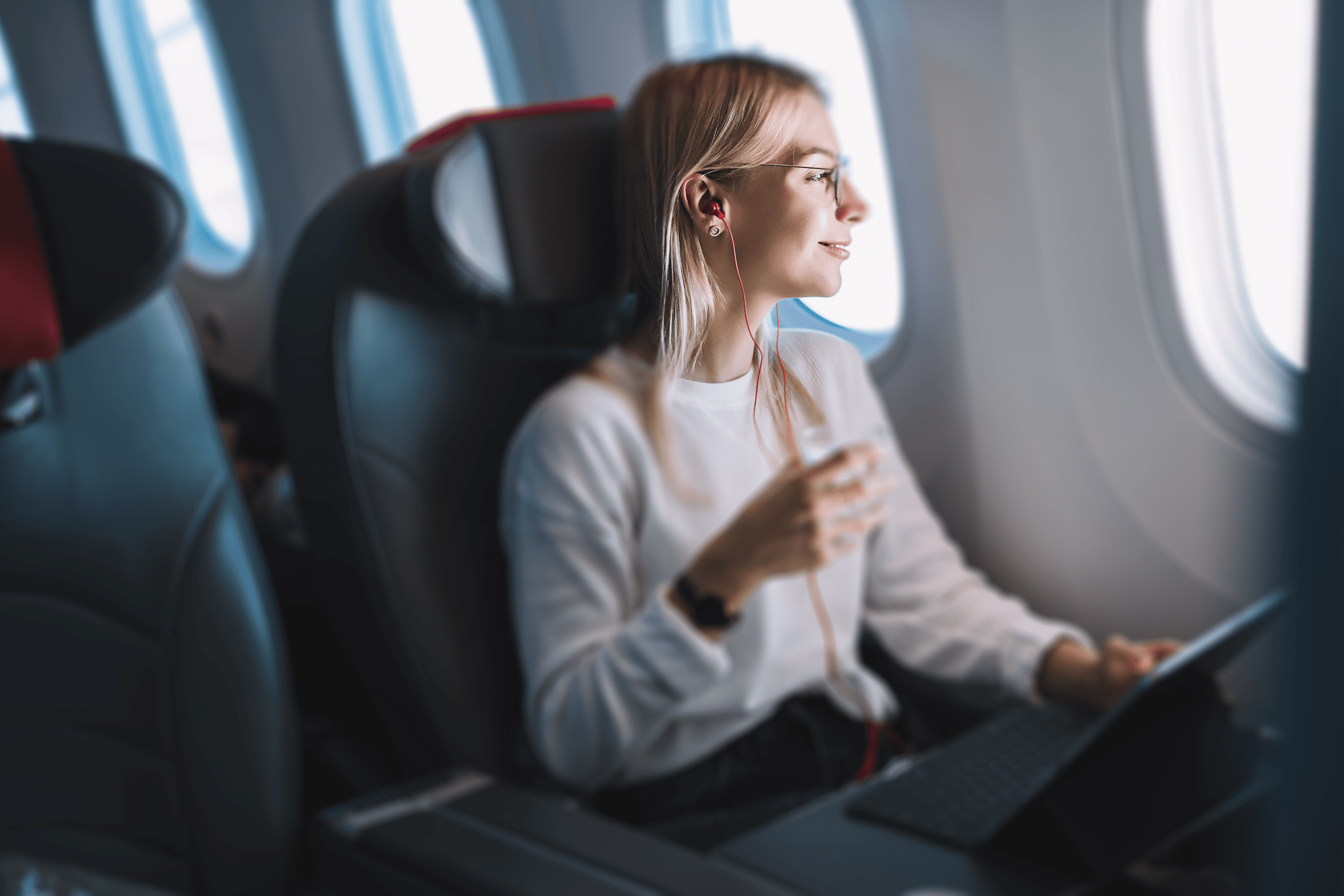 A person sits by the window on an airplane, holding a glass of water and looking out with a contemplative expression. They wear a white sweater and glasses, listening to music through earphones while using a laptop on the tray table.