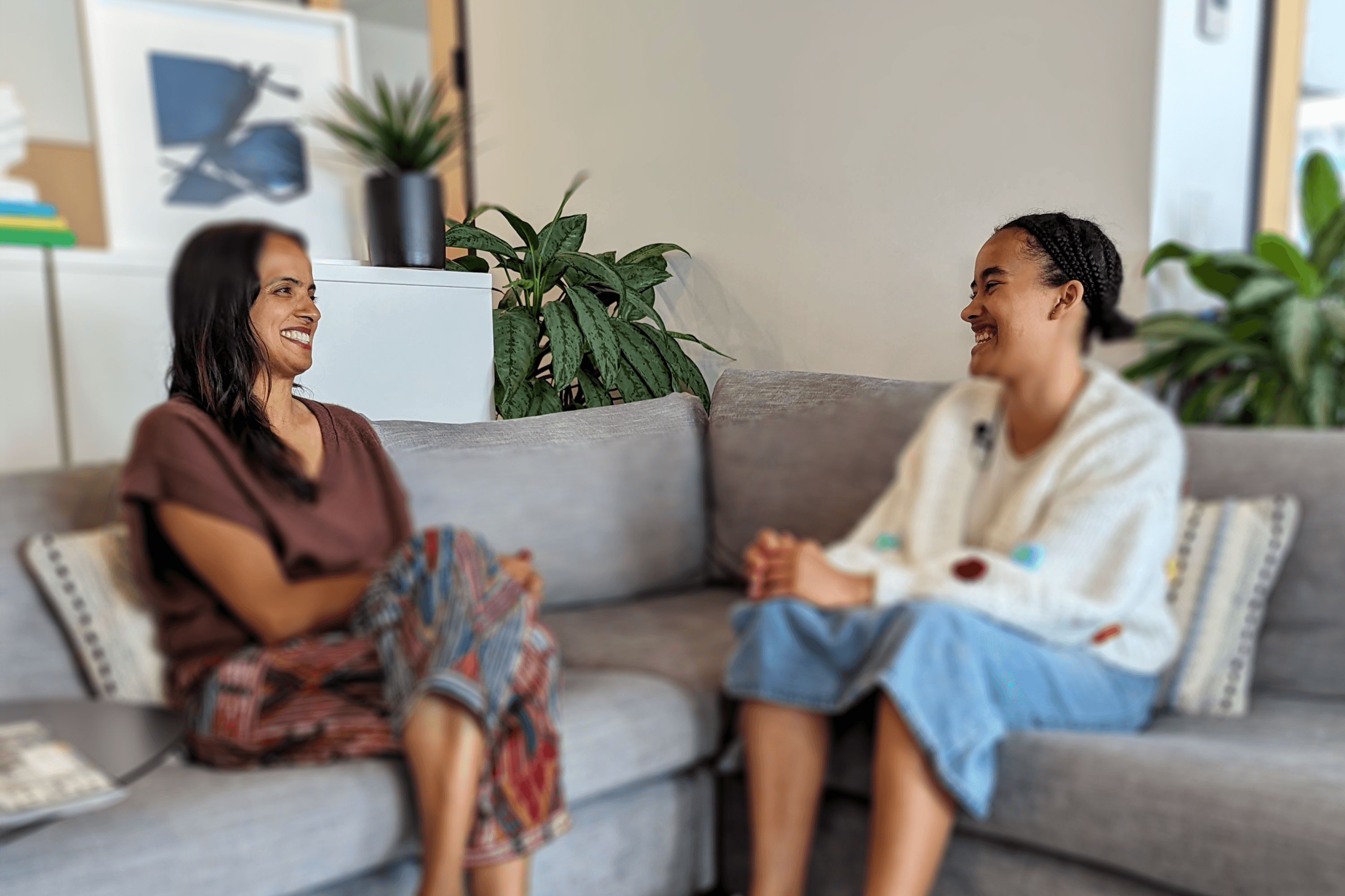 Aya Behr sits on a gray sofa with Farheen Haq. The two people are engaged in a lively conversation about unlearning. Farheen is wearing a brown top and patterned pants, laughs while sitting cross-legged. Aya, in a white cardigan with colorful patches and denim shorts, gestures animatedly.