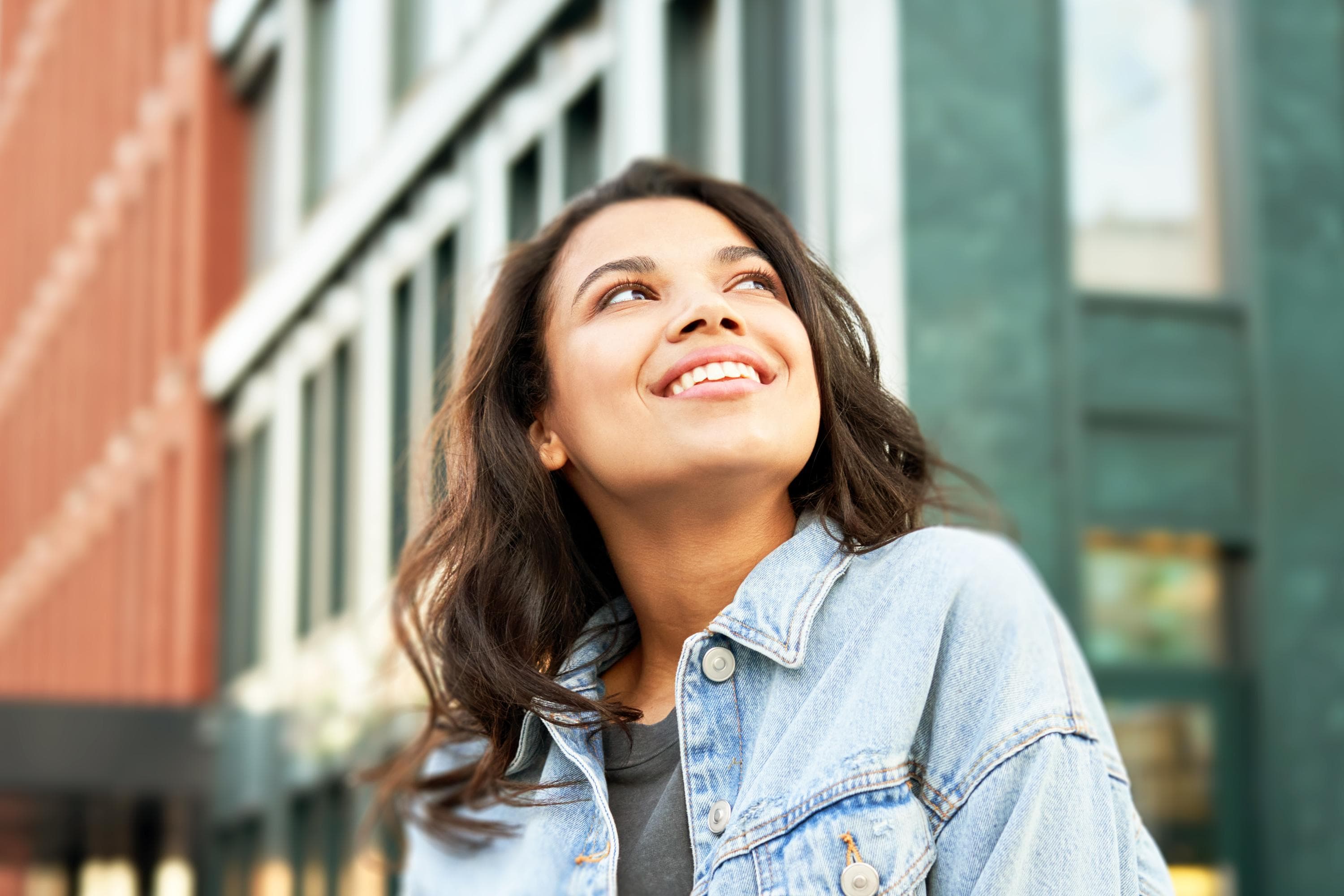 An individual wearing a denim jacket gazes upward with a joyful expression, symbolizing hope and positivity, set against an urban backdrop.