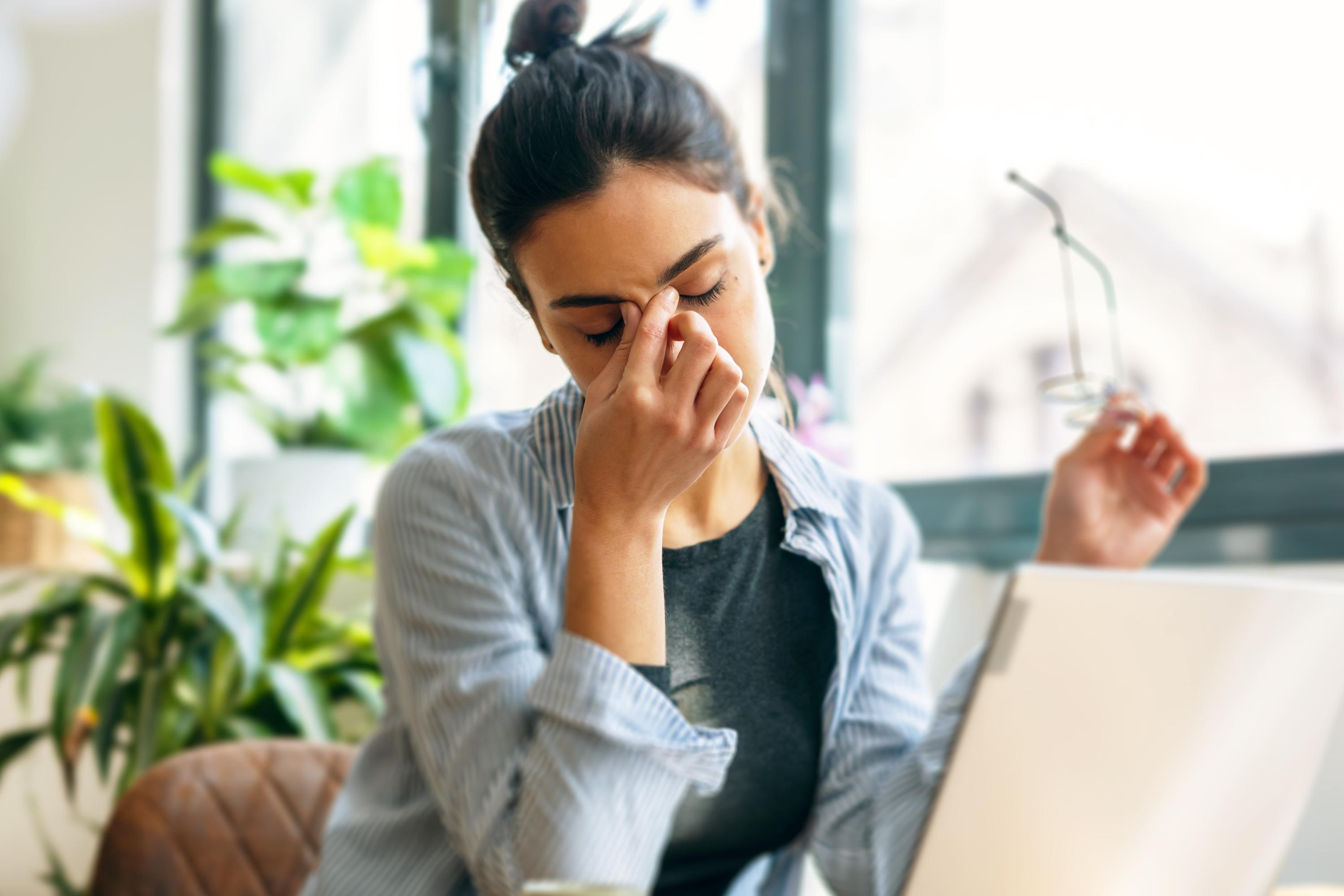 A person sitting by a window is depicted massaging the bridge of their nose, a gesture of stress or deep thought, surrounded by a serene indoor environment.