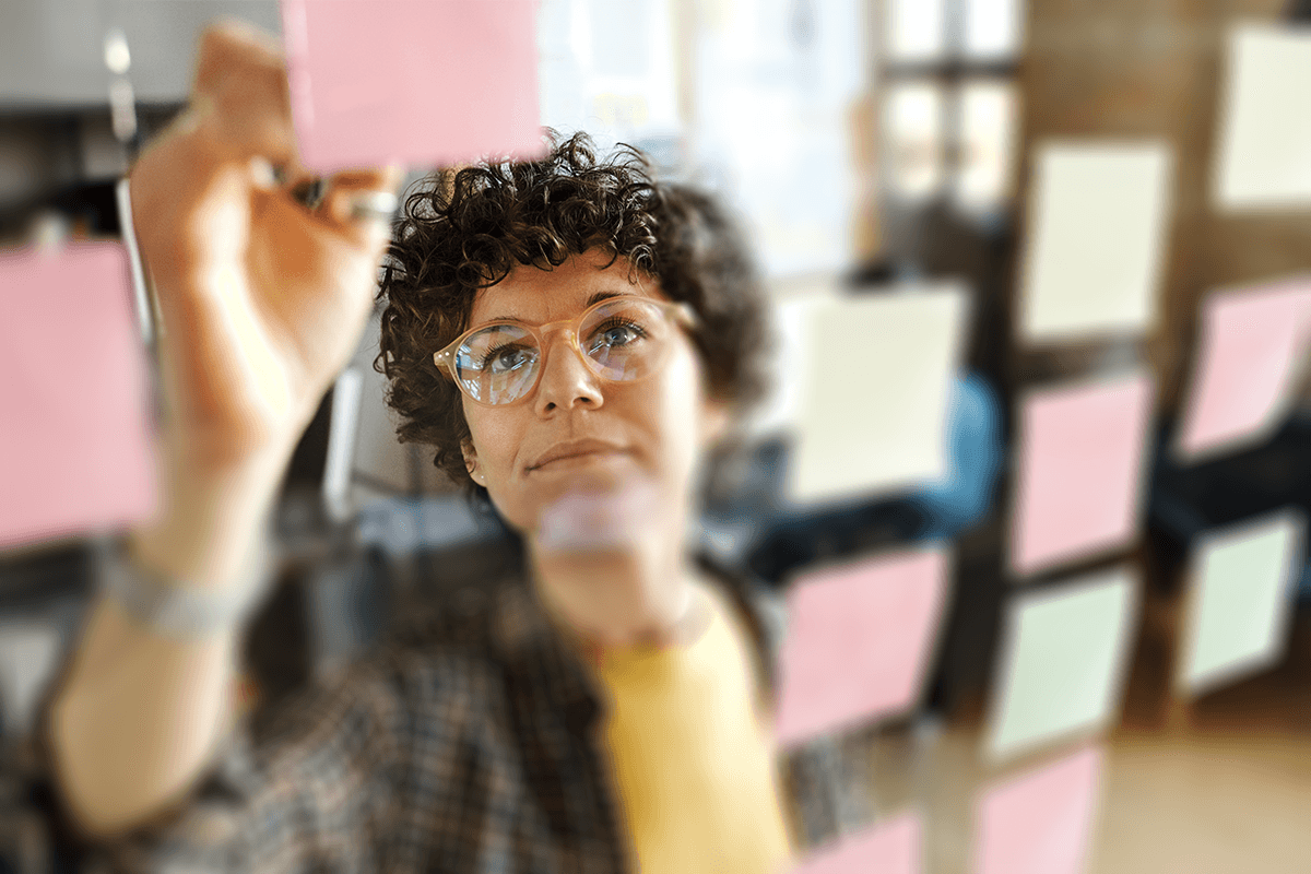 A portrait of a business founder in a modern office environment, writing on a glass wall covered in post it notes.