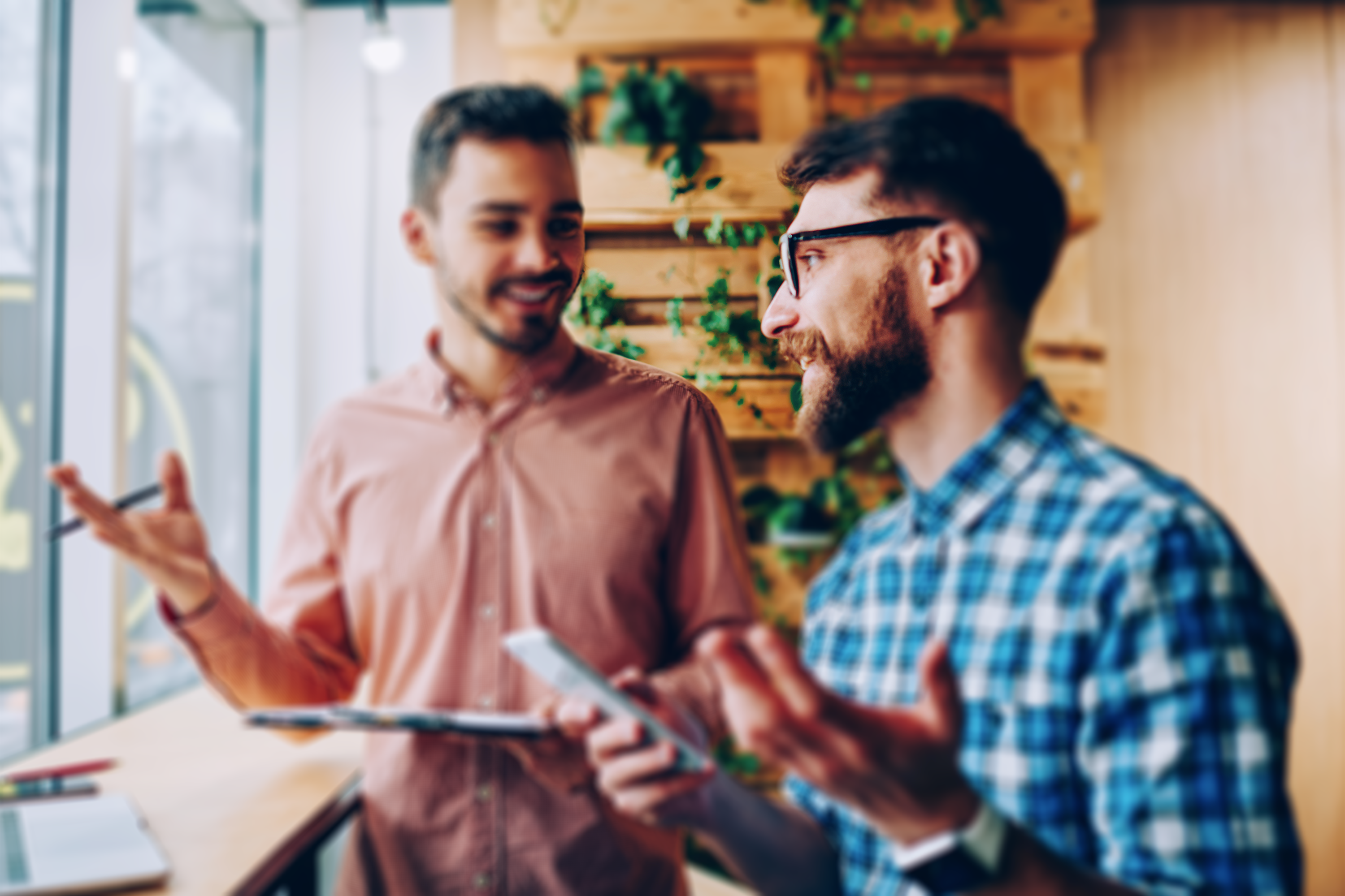 wo individuals engaging in a friendly discussion, one holding a clipboard, in a vibrant office environment with plants in the background.