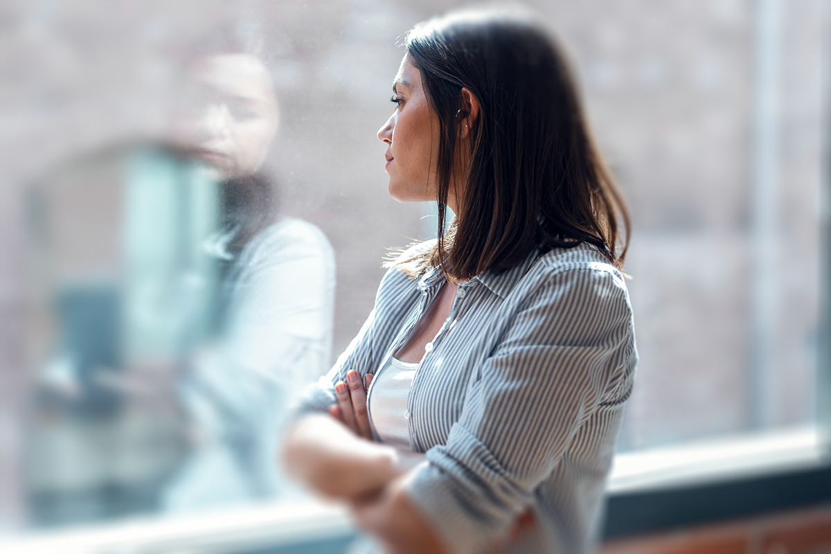 A business professional with long dark hair in a gray striped blouse stands in a modern office, arms crossed, looking through a glass window, they can see their reflection.