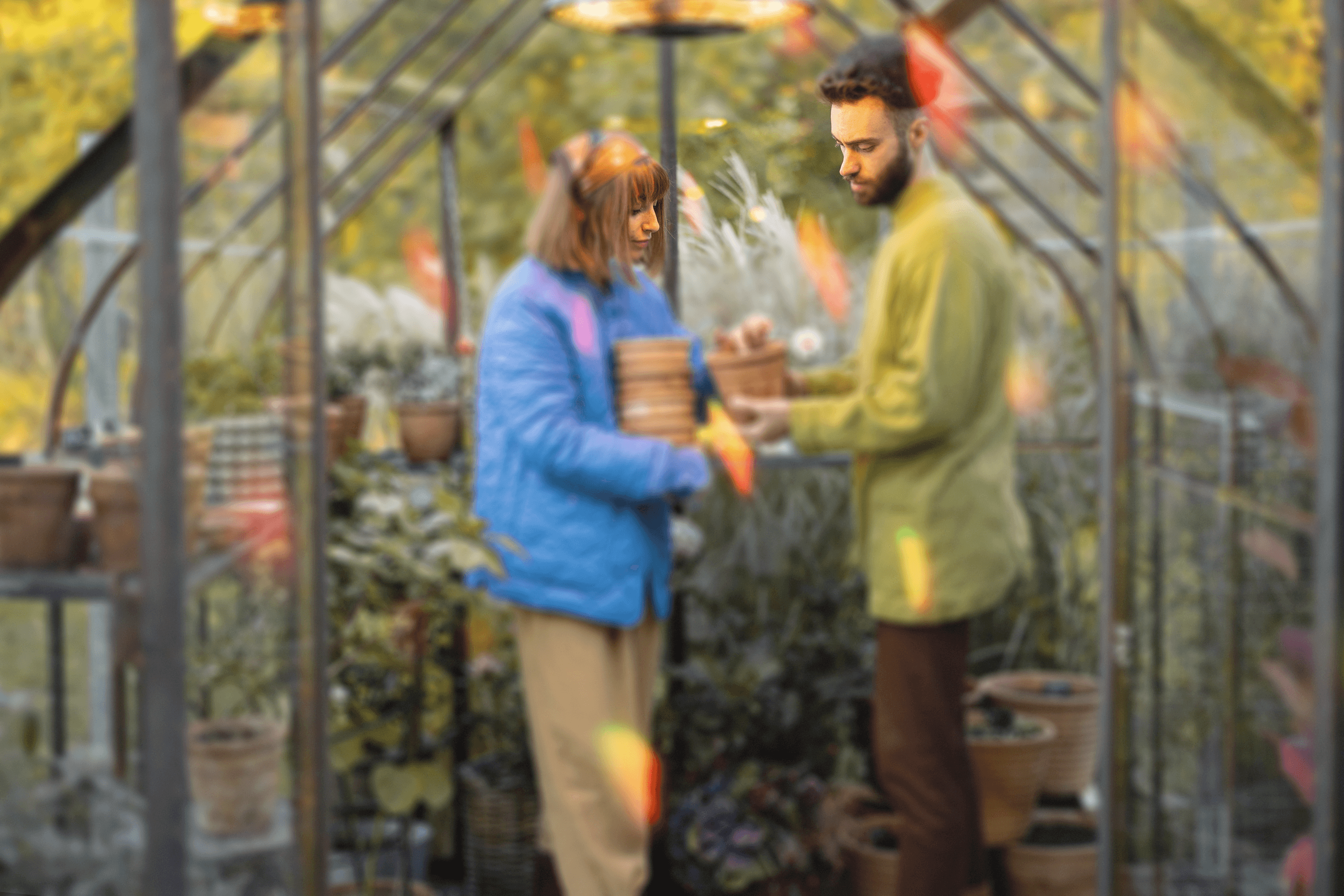 Two individuals in a greenhouse, exchanging potted plants. One, wearing a blue puffer jacket and brown hair under a headband, hands over a basket to the other, dressed in a mustard shirt and green jacket.